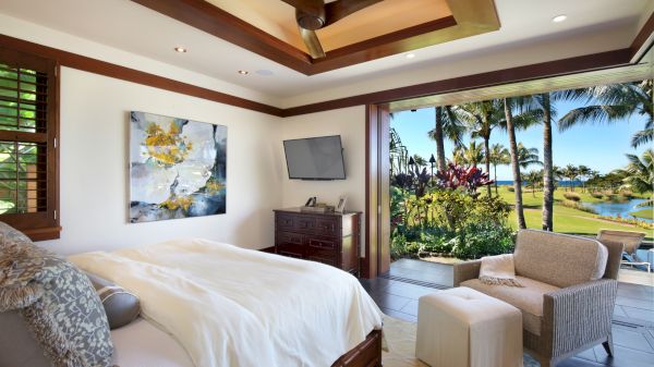 A bedroom with a bed, armchair, and TV, opens to a patio with palm trees and a scenic view. A modern ceiling fan and art piece are visible.