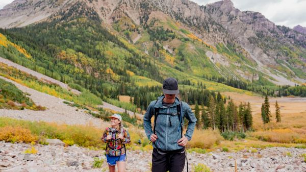 Two people in hiking gear walk in a mountainous landscape, featuring colorful foliage and rocky terrain under a cloudy sky.