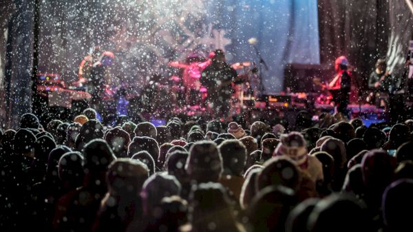 A crowd watches a band performing on stage in snowy conditions, with colorful lights illuminating the scene.