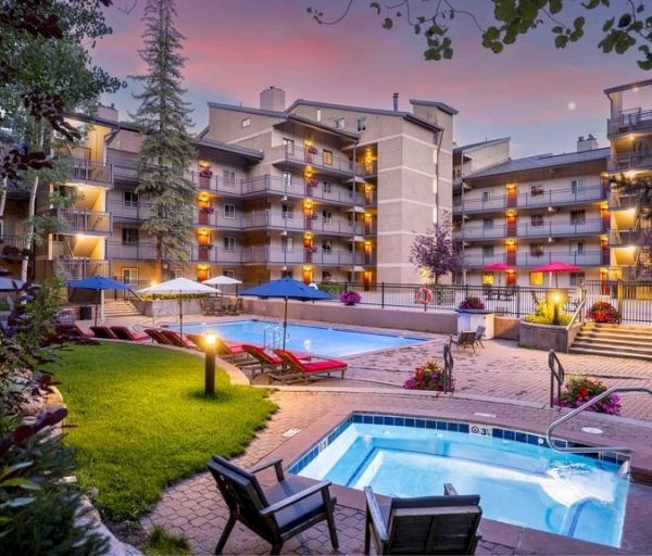 A modern hotel or resort at sunset featuring a pool, hot tub, lawn with loungers, and multi-story buildings with lit balconies.