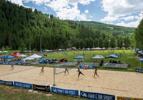 The image shows a beach volleyball game happening outdoors, surrounded by a lush green landscape and tents.
