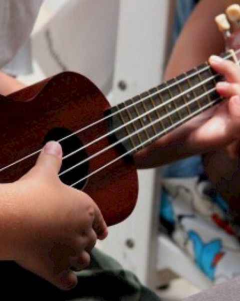 Two people are holding a ukulele, one playing it while the other assists, in a casual indoor setting.