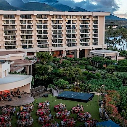 A hotel by the ocean with a lush garden, outdoor seating, and a gathering area under a clear sky with mountains in the background.
