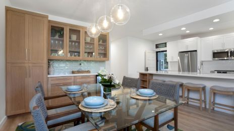 A modern dining and kitchen area features a glass table, blue dishes, wooden cabinets, bar stools, and pendant lights above.