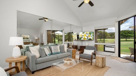 The image shows a cozy living room with modern decor, a gray sofa, large mirrors, TV, and a view of a lush green landscape outside.