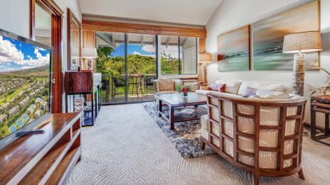 A cozy living room features a large TV, elegant sofas, and a scenic view through sliding glass doors leading to a balcony.