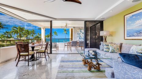 A spacious, modern living area with ocean view, featuring a dining set, sofa, glass table, ceiling fan, and art on the walls.