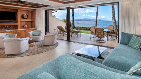 Spacious living room with blue couches, open to a patio with seating. Ocean view with palm trees outside. Cozy and inviting atmosphere.