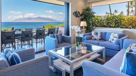 A stylish living room with blue sofas, a glass table, and a view of the ocean and mountains through large windows and an open patio area.