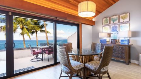 A dining area overlooks a terrace with ocean views, featuring palm trees, a table with chairs, and framed artwork on the wall.