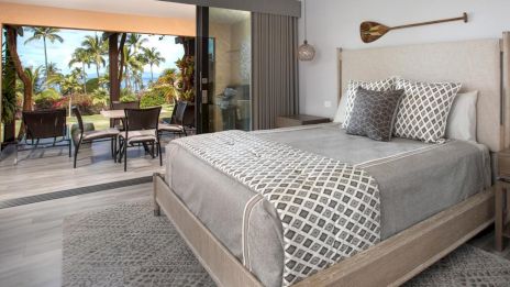A stylish bedroom with a large bed, patterned bedding, a rug, and an outdoor seating area overlooking a tropical view is shown.
