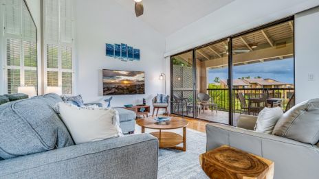 A modern living room with gray sofas, a wooden coffee table, and large sliding doors leading to a balcony with outdoor seating and scenic views.