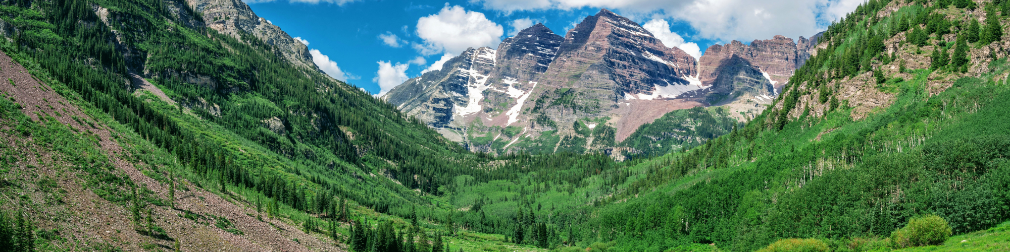 A scenic view of lush green mountains and valleys with jagged peaks under a bright blue sky dotted with clouds, showing natural beauty.