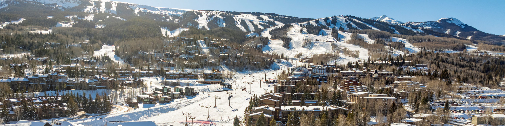 A ski resort landscape with snowy slopes, ski lifts, and cabins nestled in a forested mountain area under a clear blue sky.