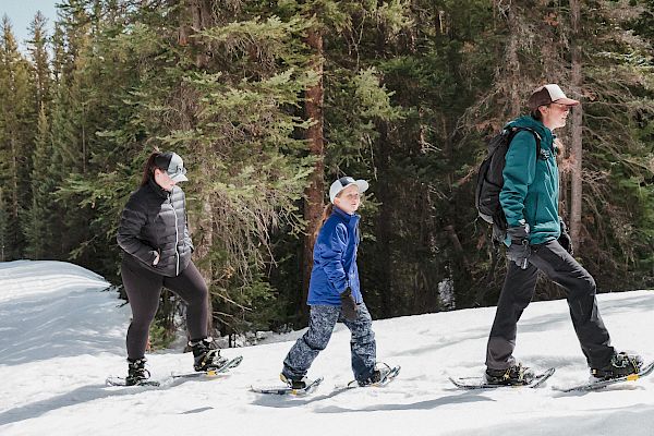 Three people are snowshoeing on a snowy trail through a forested area, dressed warmly for cold weather.