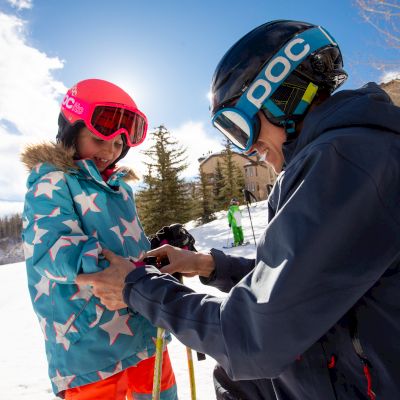A person in ski gear is helping a child adjust clothing on a snowy slope, with a ski lift and trees in the background.