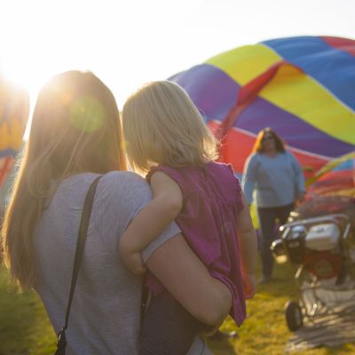 A woman holds a child while standing near colorful hot air balloons and other people in a sunny outdoor setting.