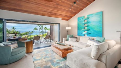 A modern living room with a wooden ceiling, white sofas, and blue accents, opens to a patio with palm trees and ocean view.