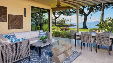 A stylish outdoor patio with a sofa, modern furniture, a blue rug, and a dining table, overlooking a garden and ocean view.