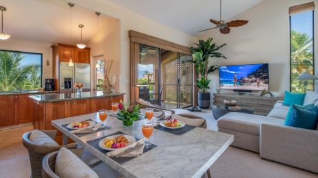 A modern open living space with a kitchen, dining table set for breakfast, and a lounge area with a TV and tropical decor.