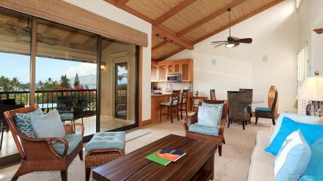 A cozy living room with wicker furniture, blue accents, a wooden table, and a view of a balcony with lush greenery and clear skies.