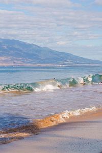 The image features a serene beach scene with gentle waves, sandy shore, and distant mountains under a partly cloudy sky.