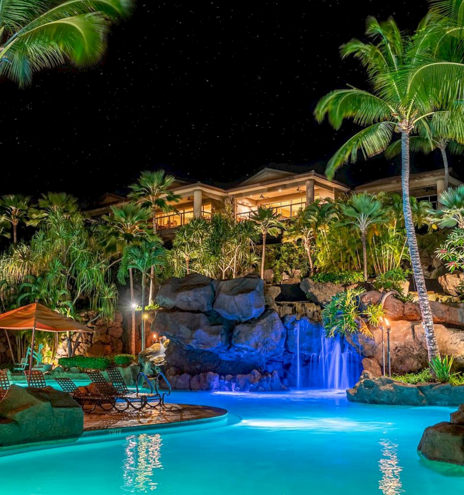 A tropical resort scene with a pool, waterfall, lush plants, and lounge chairs under umbrellas at night.
