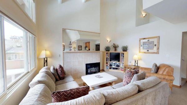 A cozy living room with a sectional sofa, fireplace, TV, and lamps. Large windows allow ample natural light.