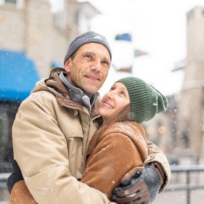 A couple in winter clothing embraces outdoors with a snowy backdrop, smiling and looking content.