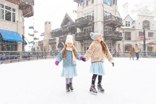 Two girls in winter attire are ice skating outdoors, holding hands, with snow falling around them in a charming town setting.