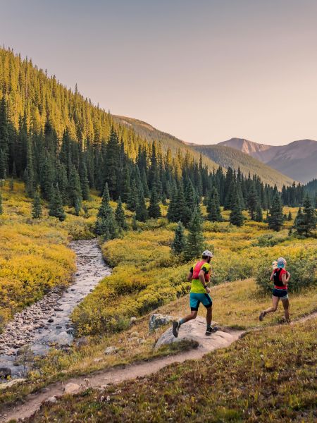 Two people and a dog run along a trail in a picturesque forested valley, beside a stream under a clear sky.