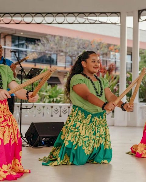 Three young dancers in colorful skirts perform with sticks, accompanied by a musician on stage in an outdoor setting.