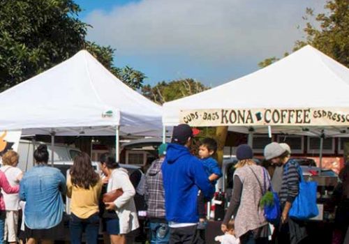 People are gathered around tents at an outdoor market or festival, with one tent labeled for Kona coffee.