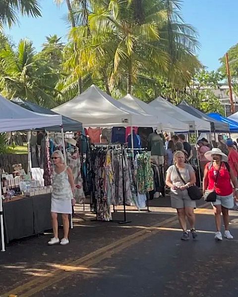A street market with people browsing stalls selling clothes and various items, under tents with palm trees lining the road.