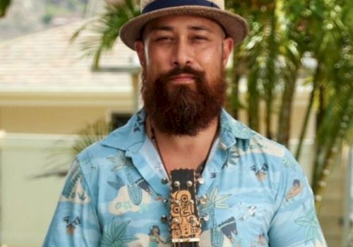 A person with a beard, wearing a hat and a tropical shirt, stands with a string instrument outdoors near palm trees.