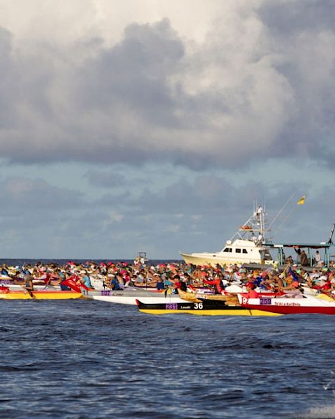 The image depicts a group of colorful canoes racing on the ocean with several boats in the background under a cloudy sky.