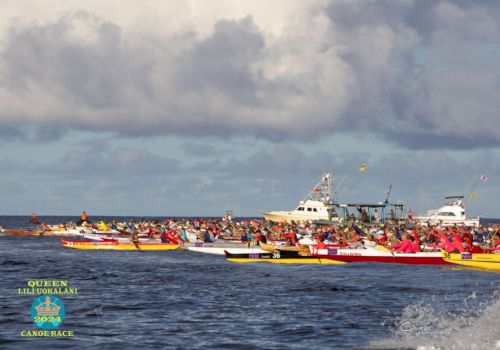The image depicts a group of colorful canoes racing on the ocean with several boats in the background under a cloudy sky.