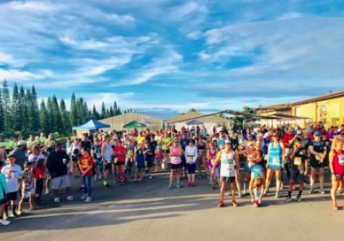 A large group of people, likely participating in a running event, gathers outdoors with a backdrop of trees and clear skies.