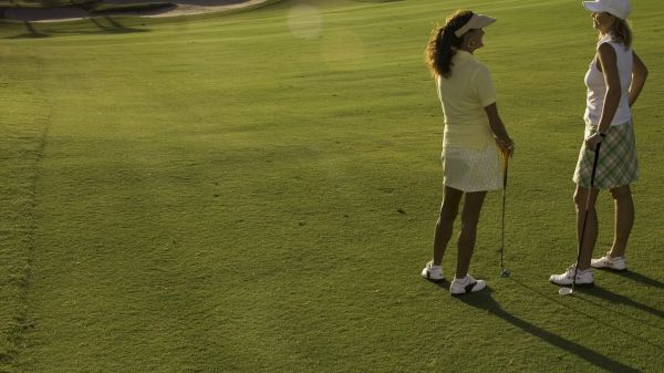Two individuals are standing on a sunny golf course, holding clubs and wearing golf attire.