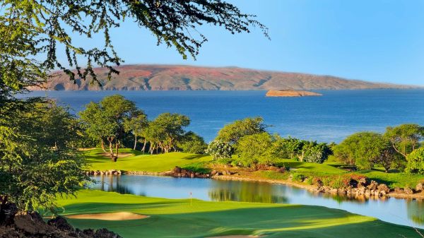 A scenic view of a golf course by the ocean, with lush greenery, a small island in the distance, and blue skies overhead.