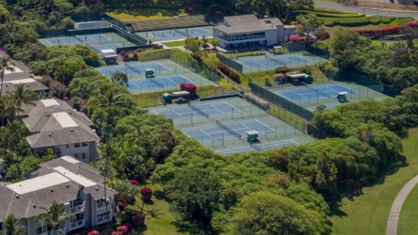 The image shows an aerial view of several tennis courts surrounded by greenery, buildings, and a parking area in a scenic landscape.