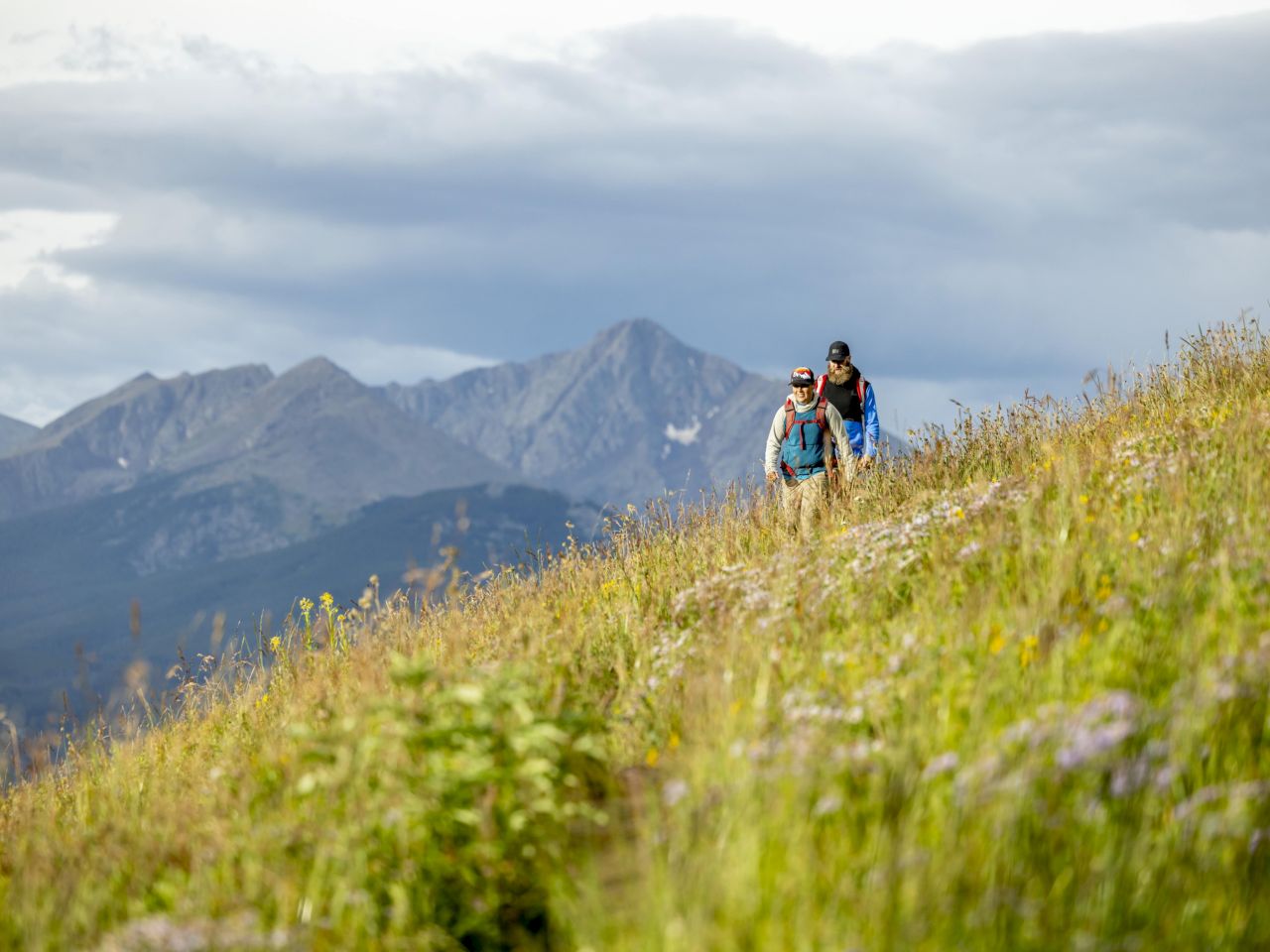 Two hikers navigate a grassy hillside against a backdrop of mountains under a cloudy sky. Nature's beauty surrounds them.