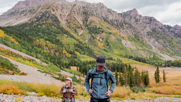 The image shows two hikers in a colorful mountain landscape, with vibrant green trees and rocky terrain in the background.