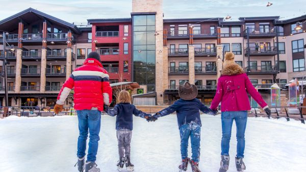 A group of four, including kids, ice skating and holding hands in front of a modern building with snowy surroundings.