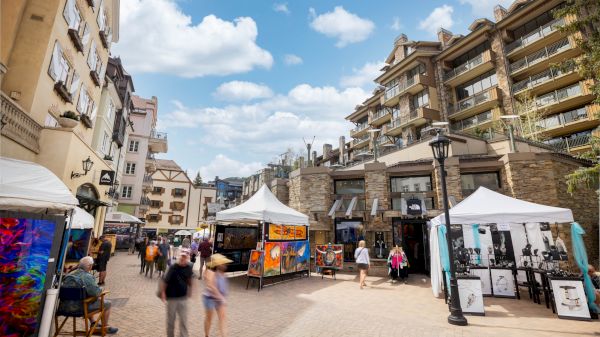 A lively street scene featuring art stalls, shops, and people enjoying their day under a partly cloudy sky.