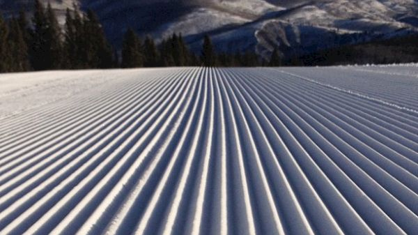 The image shows snow-covered slopes with groomed lines and mountains in the background against a clear blue sky.