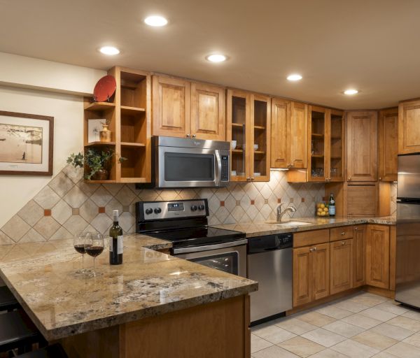 The image shows a cozy kitchen featuring wooden cabinets, stainless steel appliances, granite countertops, and a wine setup.