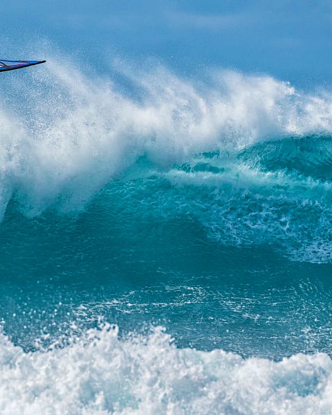 A windsurfer performs an acrobatic jump over vibrant blue waves, captured in a moment of thrilling action. The ocean sparkles.