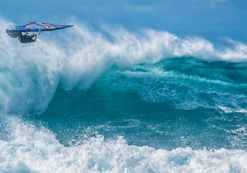 A windsurfer performs an acrobatic jump over vibrant blue waves, captured in a moment of thrilling action. The ocean sparkles.