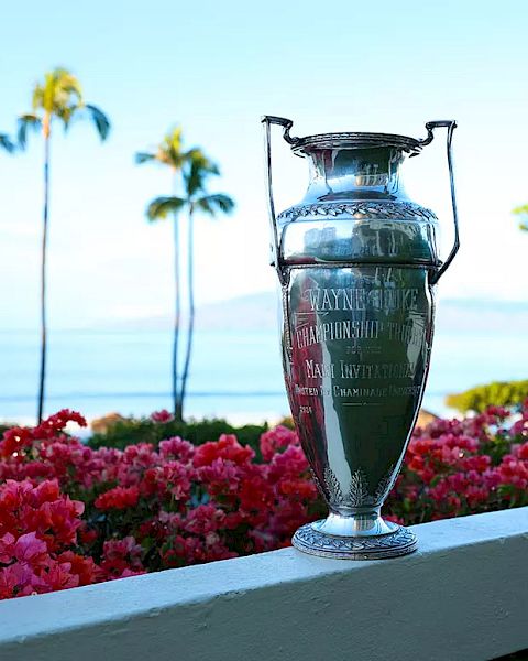 The image features a trophy resting on a ledge, surrounded by vibrant pink flowers, with a scenic ocean view and palm trees.